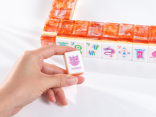 Coral Lotus American Mah Jongg Joker tile held in a hand beside an orange tile wall