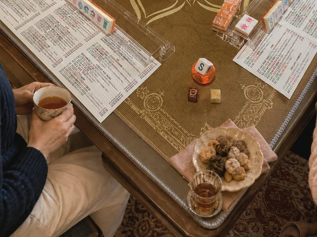 Hands holding a teacup beside snacks, a drink and mah jongg tiles at the corner of a game table